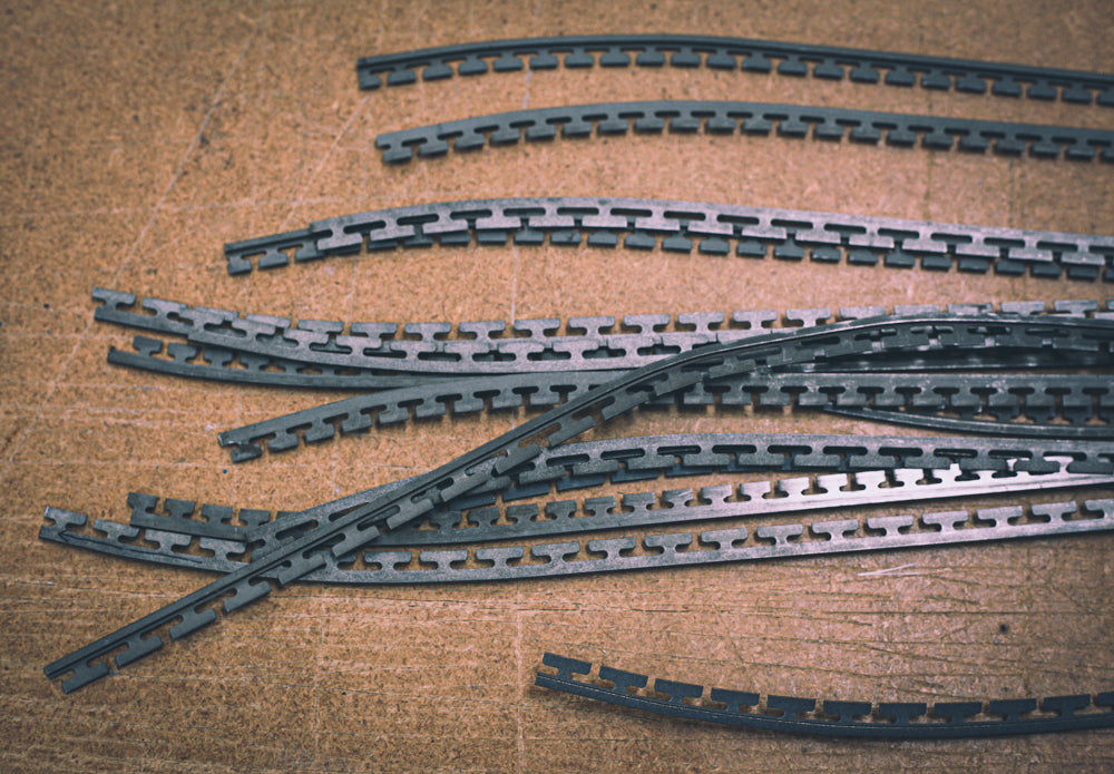 A close up image of raw metal edges that have been bent into the shape of a snowboard before they’ve been glued to the base of the board. There are metal teeth on one side of the edges and a straight metal edge on the other side.