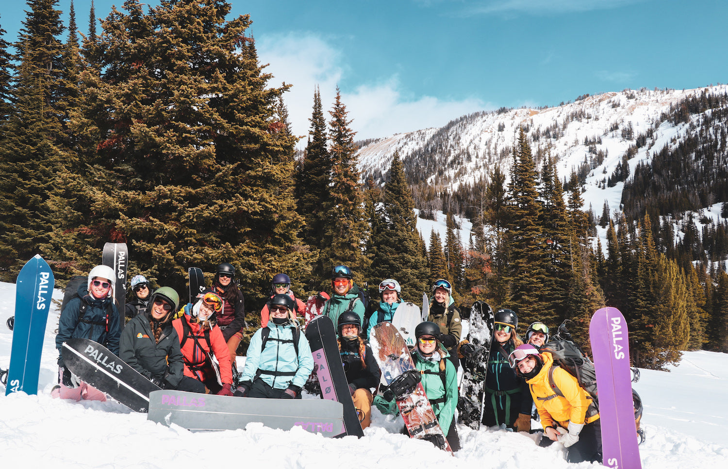 Image of 15 women sitting in the snow, smiling at the camera. It’s a bright day, with blue sky and many women wearing sunglasses. They are on a women’s splitboard tour with many of them demoing Pallas splitboards.