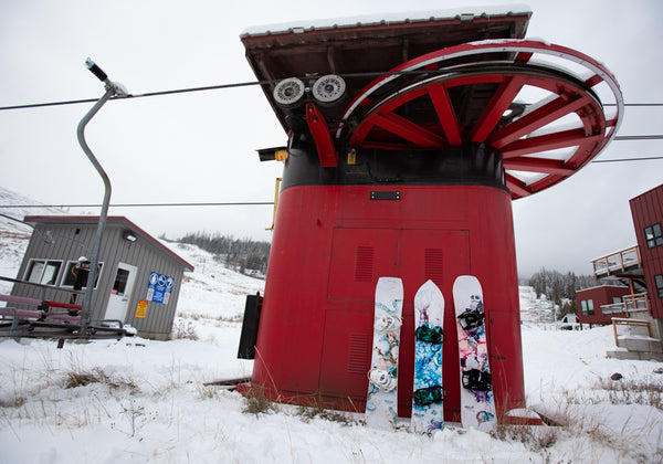 Three Pallas snowboards are leaned up against the base terminal of a red chairlift. The chairlift is an old, fixed-grip, two-seater.