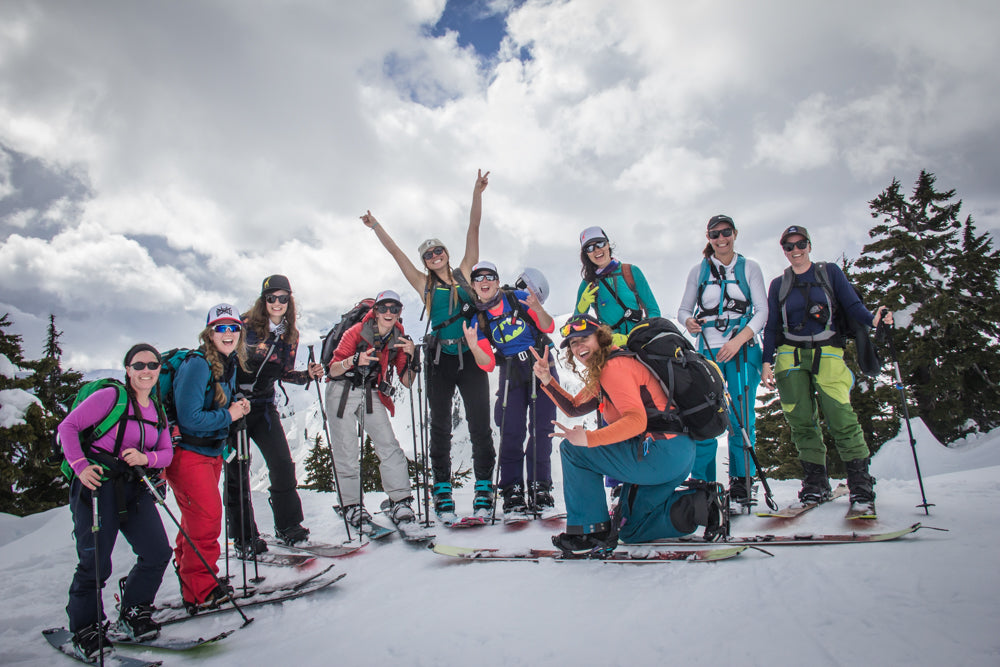 Image of 10 women standing on a ridgeline and stopping to take a group photo during a splitboard tour. The sky is cloudy but they are all wearing sunglasses and hats. They are all smiling at the camera and some are throwing up peace signs.