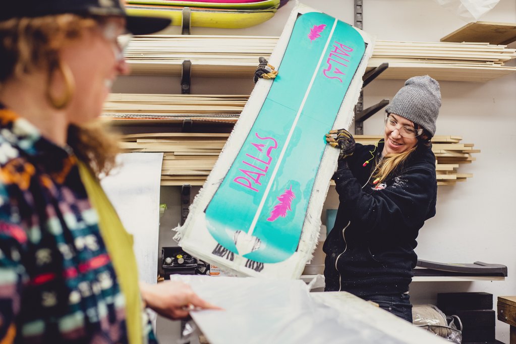 An image of a woman wearing eye protection, gloves and ear plugs. She is holding a Pallas splitboard that is fresh out of the snowboard press. Fiberglass and epoxy cover the base of the board, which is pointing towards the camera.