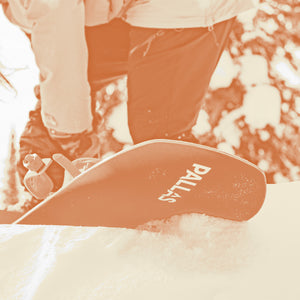 Image of a snowboarder strapping into their bindings on their Pallas Hedonist snowboard before descending. The tail of the board is above the snow, showing the Pallas logo on the base.