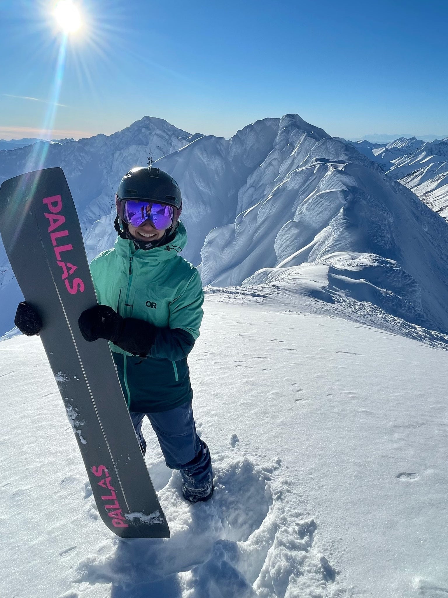 A snowboarder stands atop a snowy, alpine mountain on a bluebird day. The sun is shining behind her, and she's holding a Pallas Epiphany splitboard for the camera.