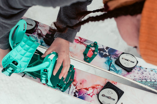 A woman is bending over the Pallas Hedonist splitboard and attaching softboot compatible splitboard bindings from Spark onto the board.