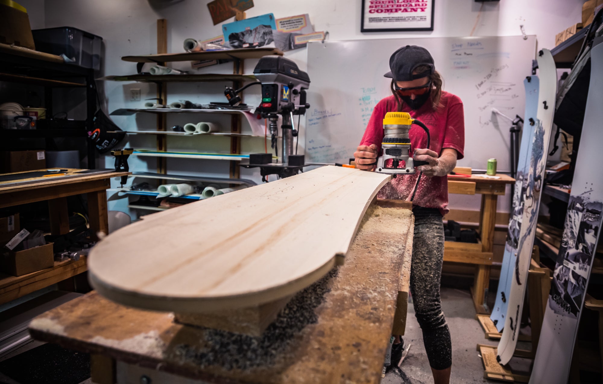 Image of a woman wearing eye protection and focused on using a router tool in the Pallas Snowboards workshop. They are cutting a wood core to the specific shape and length. The woman and the workbench are covered in wood shavings.