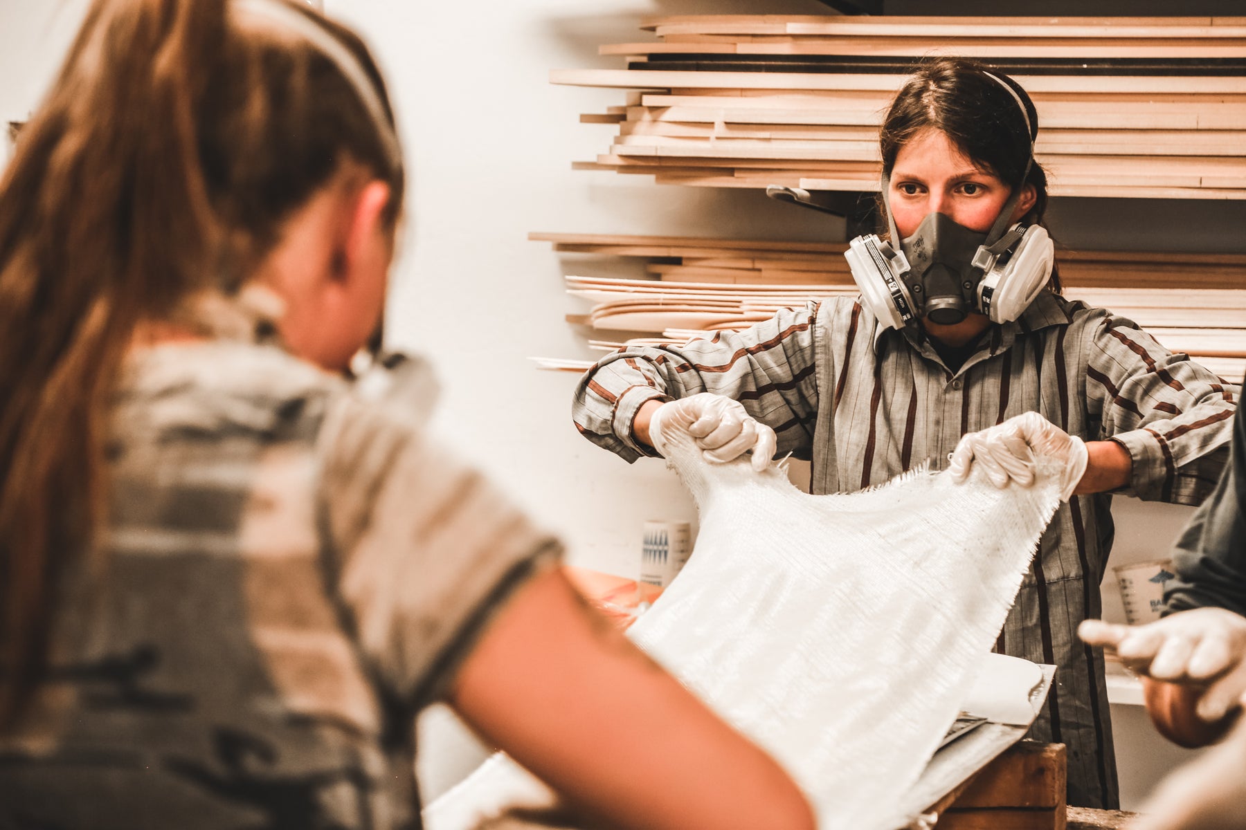 Two women wearing respirator masks and laying down a piece of epoxy-covered fiberglass onto a snowboard being built at the Pallas Snowboards workshop in Salt Lake City, Utah.