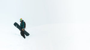 Image of a woman standing on top of a snowy hill dressed in winter snowboard gear, holding her Pallas Epiphany snowboard and looking at the camera. 