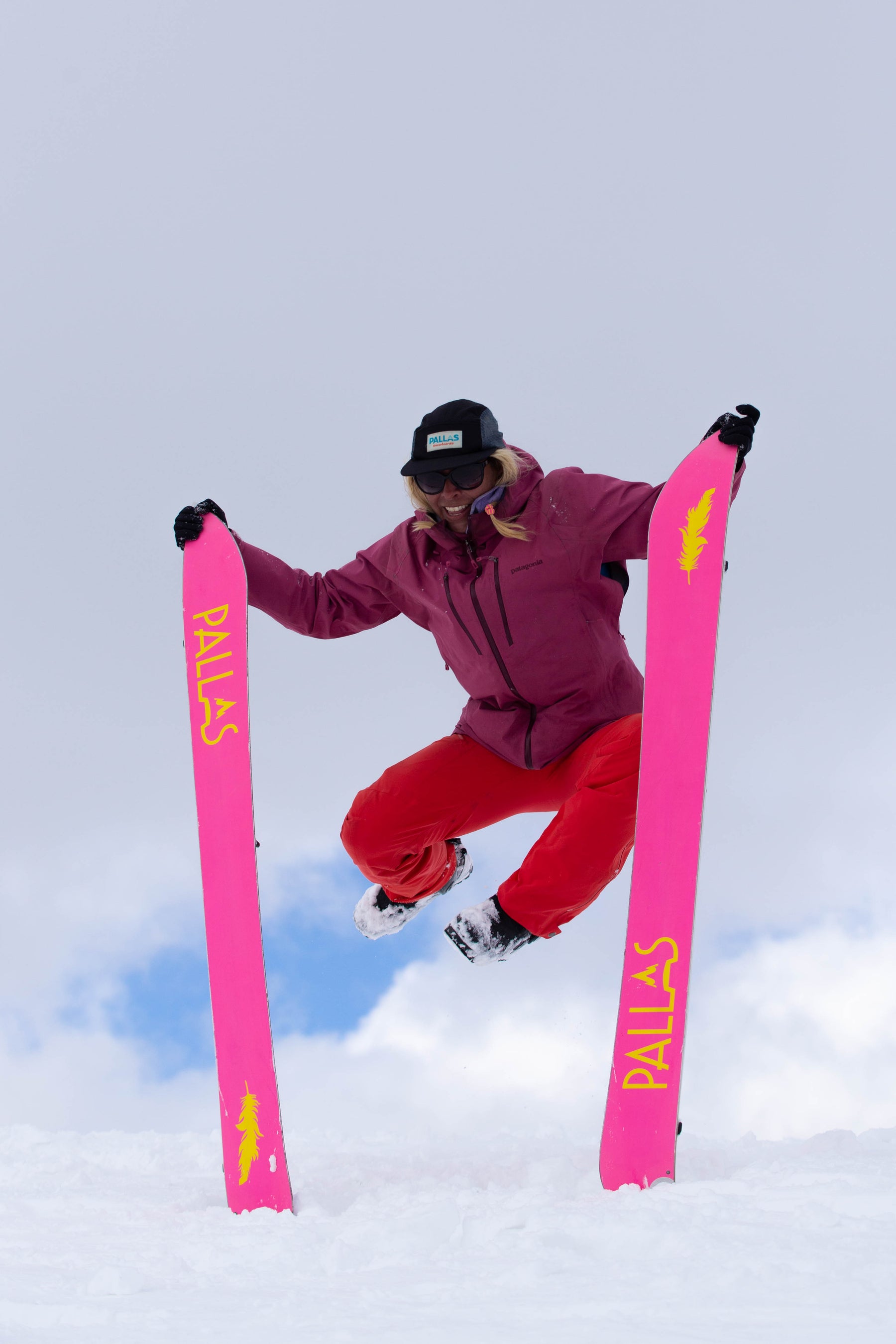 A woman is holding the skis of her Pallas splitboards upright in the snow while she jumps in the air. The base of the skis are bright pink and the woman is smiling in the air.