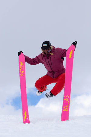 A woman is holding the skis of her Pallas splitboards upright in the snow while she jumps in the air. The base of the skis are bright pink and the woman is smiling in the air.