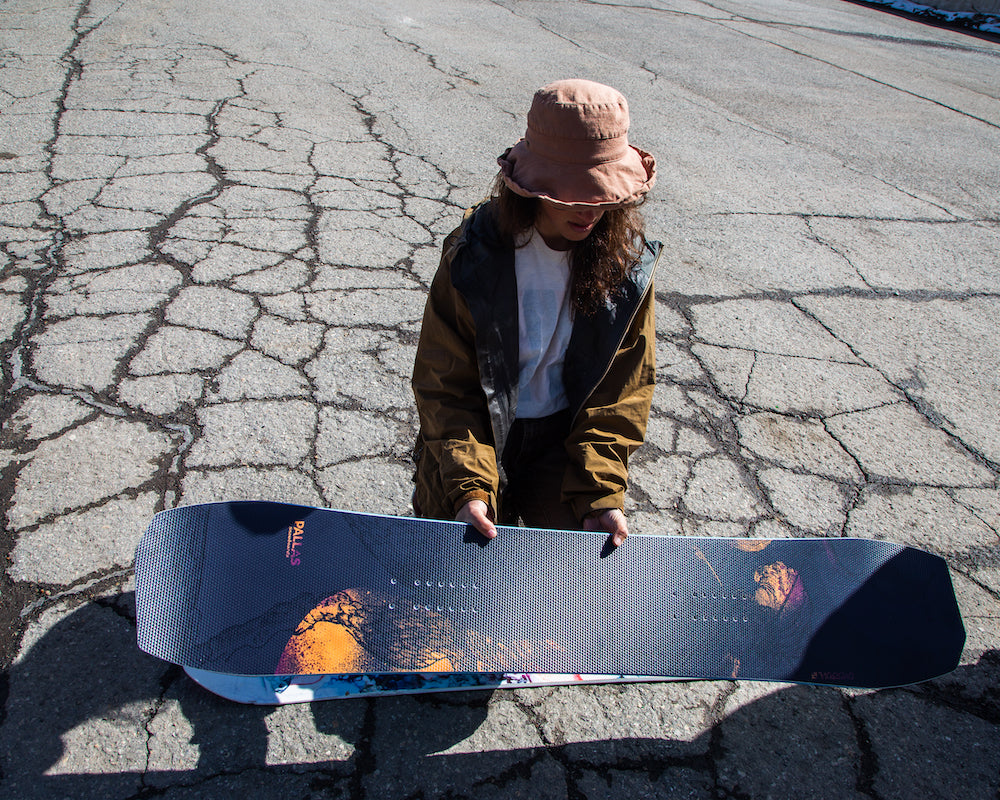 A woman holds the Pallas Oversoul snowboard with powder inserts in a parking lot.