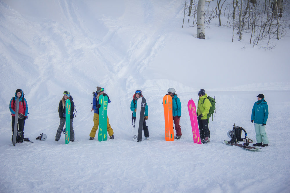 Seven splitboarders are standing at the base of a run, holding their splitboards upright and showing the colorful bases of all the boards.